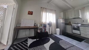 Quarto compacto com cama, escrivaninha, ventilador, pequeno espaço de cozinha e janela com cortinas.