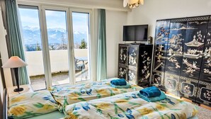 A hotel room with two beds, a TV, a decorative cabinet, and a view of mountains through large windows. - Vermala Soleil B 13 COMBI by Interhome (Crans-Montana)