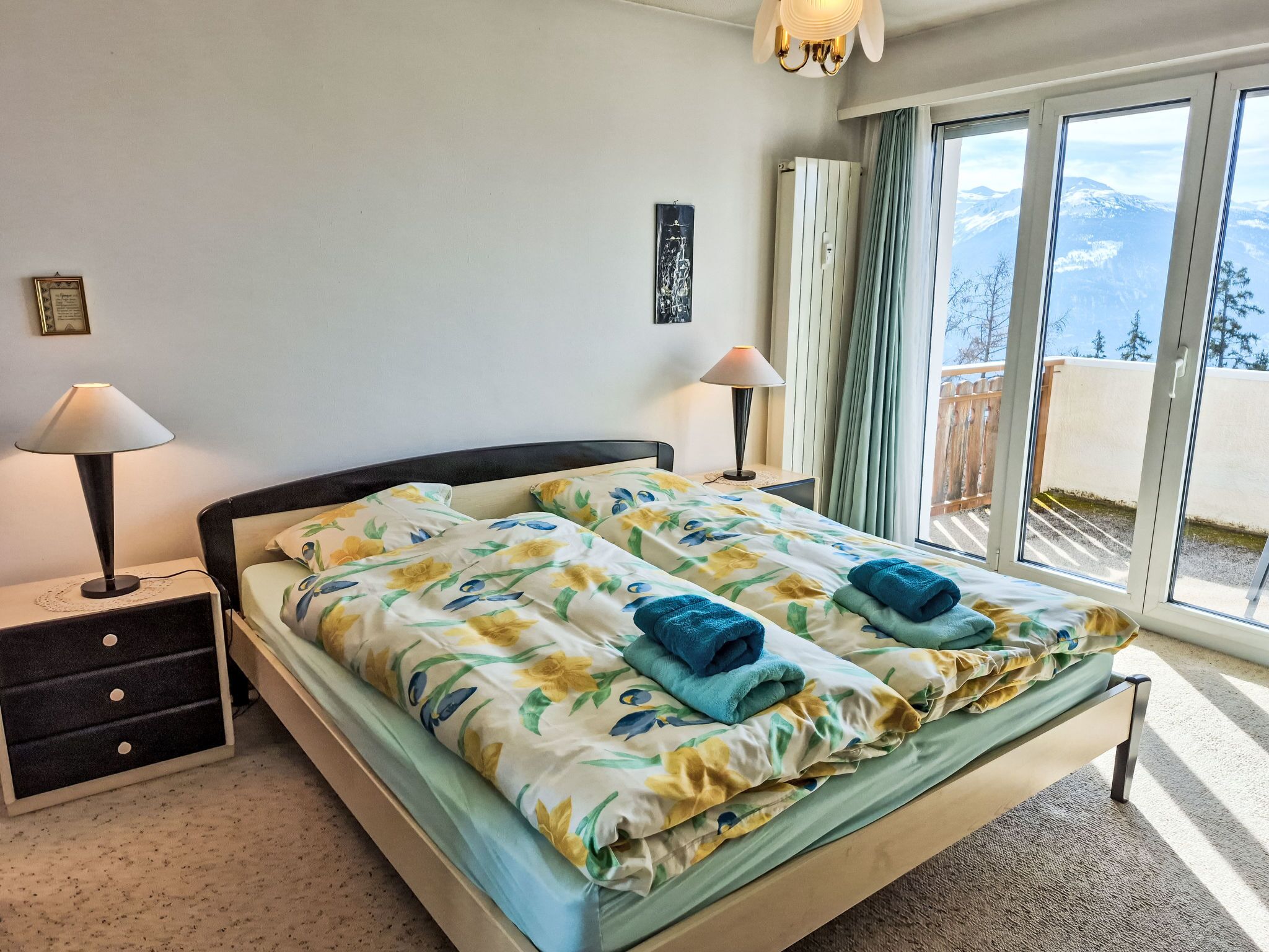 A bedroom with a bed, two bedside lamps, a nightstand, a window with a view of mountains, and a ceiling light.
