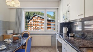 A modern kitchen with a large window, a dining area with a table and chairs, and a view of a multi-story building. - Vermala Soleil B 13 COMBI by Interhome (Crans-Montana)