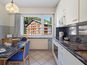 Eine moderne Küche mit großem Fenster, einem Essbereich mit Tisch und Stühlen und Blick auf ein mehrgeschossiges Gebäude.