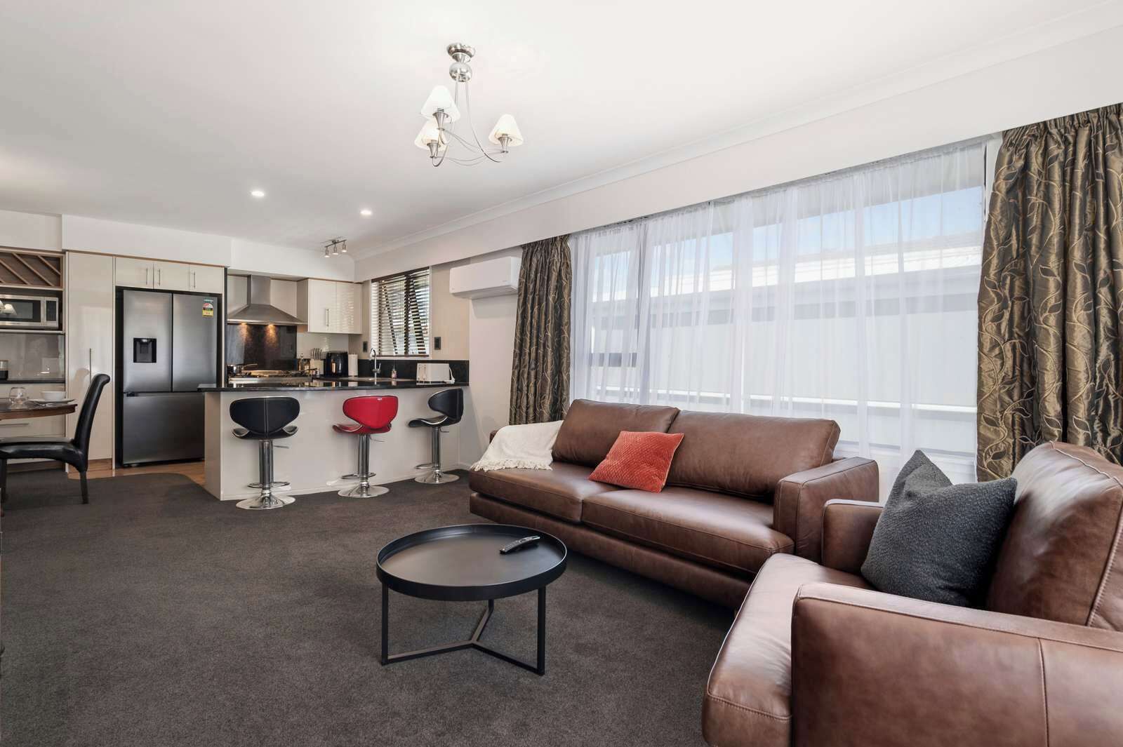 A modern living room with a brown leather sofa, a small black coffee table, and a kitchen area with a refrigerator and bar stools.