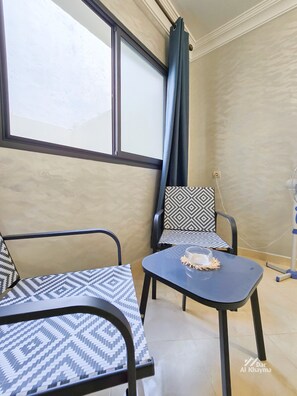 Ein Zimmer mit zwei Stühlen, einem Tisch, einem fenster mit Vorhängen und einem Wandventilator.