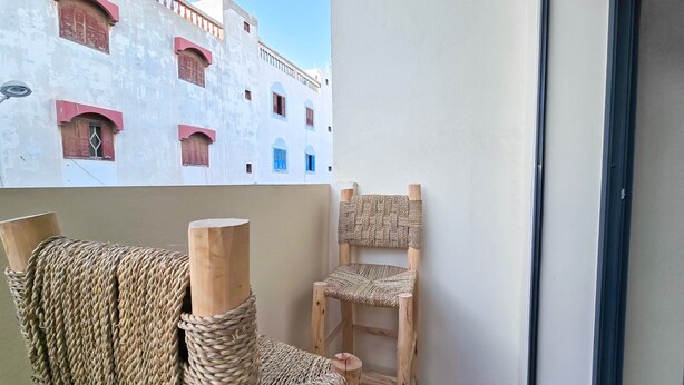 Un balcone con una sedia in vimini e una sedia di legno, un edificio bianco con infissi rossi e un cielo sereno.