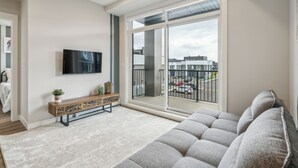 A modern living room with a grey sofa, a wooden console table with decorative items, a flat-screen TV mounted on the wall, and a large sliding glass door leading to a balcony with a city view. - Eclectic New Condo in Seton’s hub (Calgary)