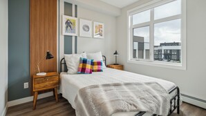 A bedroom with a bed, wooden nightstands, a window with a view, and wall-mounted artwork. - Eclectic New Condo in Seton’s hub (Calgary)