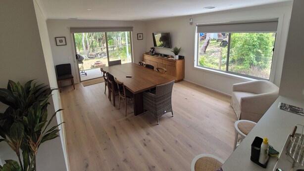 A spacious living area with a wooden dining table, a kitchen area with a counter, and large windows offering views of the outdoors. - Golden Beach Holiday Home (Golden Beach)
