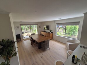 A spacious living area with a wooden dining table, a kitchen area with a counter, and large windows offering views of the outdoors.