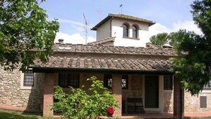 Rakennuksessa on tiilikatto, kiviseinät ja pieni puutarha, jossa on yksi puu ja punainen kukka.