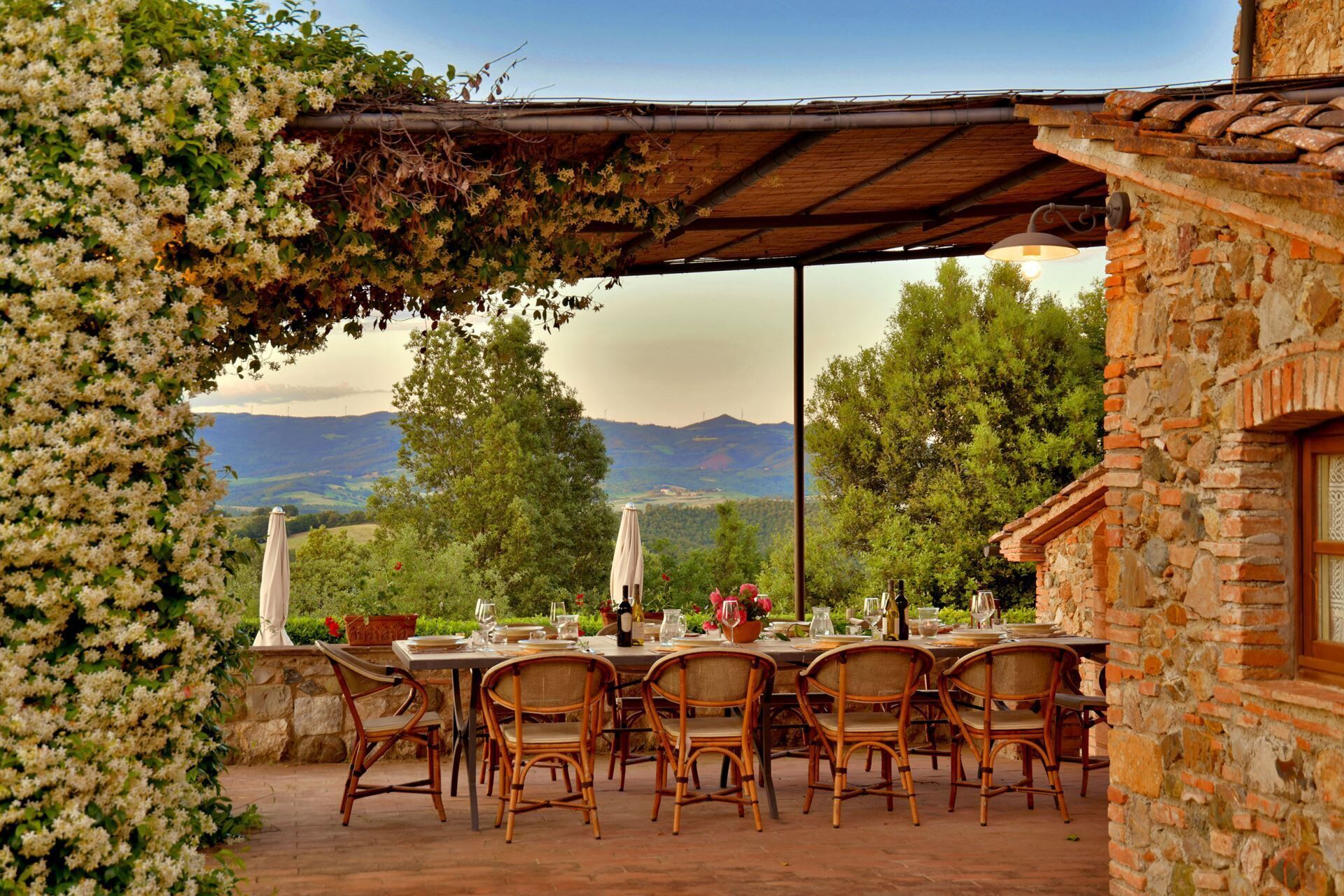 An outdoor dining area with a table set for six, surrounded by wicker chairs, under a pergola with climbing plants. A stone building with a brick facade is visible to the right.