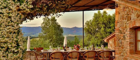 An outdoor dining area with a table set for six, surrounded by wicker chairs, under a pergola with climbing plants. A stone building with a brick facade is visible to the right.