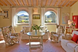 A cozy living room with floral-patterned chairs, a round table, and large windows offering scenic views. - Tuscan farmhouse in the hills 15 km from the beach. For those who looking for relax, peaceful and su (Guardistallo)