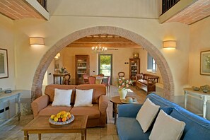 A spacious living area with a high ceiling, exposed wooden beams, and a brick archway. There are two sofas, a coffee table with fruit, and a dining area visible through the archway. - Magnificent stone house 10 km from the sea with solarium and heated pool. (Guardistallo)