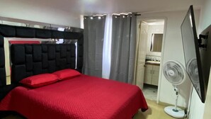 Una camera da letto con un letto, un comodino, uno specchio, una ventola e un bagno visibile attraverso una porta aperta.