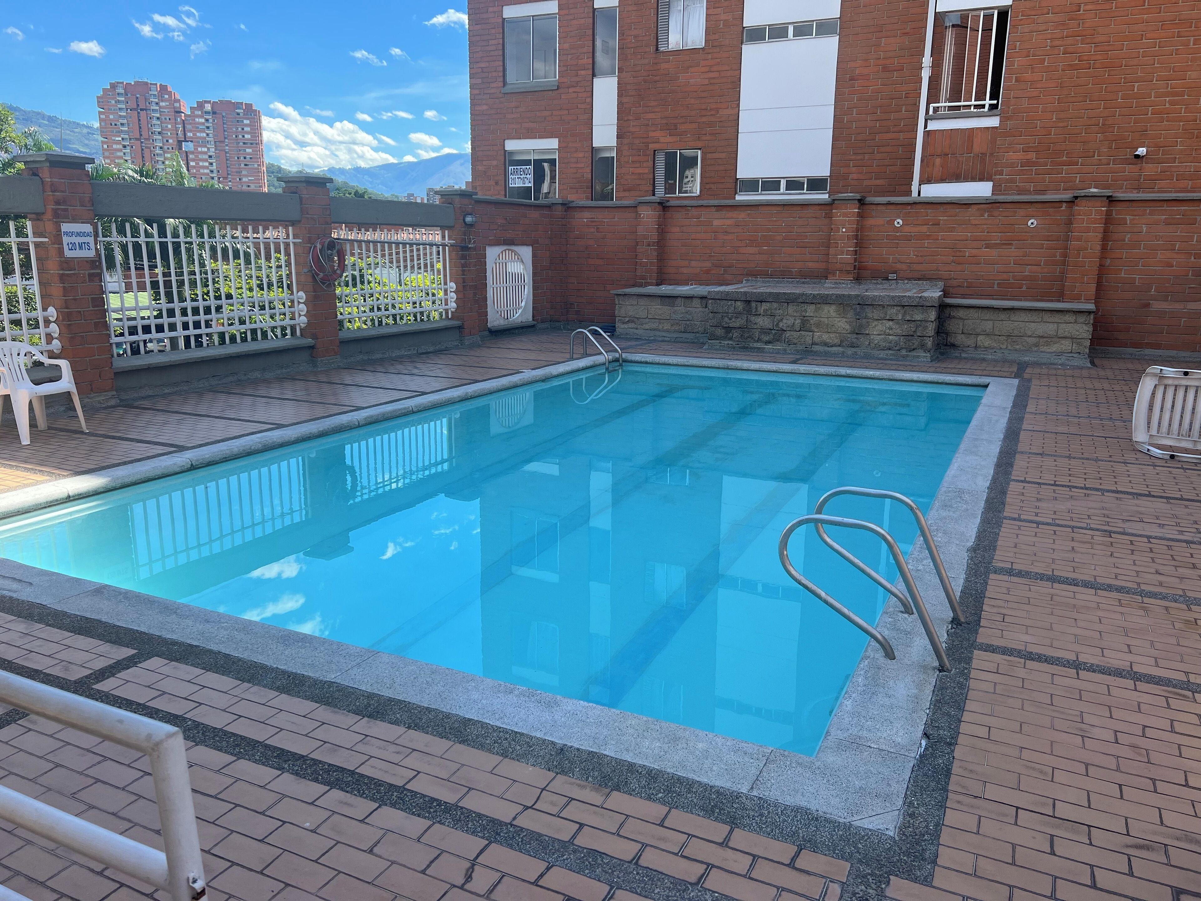 Una piscina rectangular con una escalera de metal, rodeada por un patio de ladrillo con valla blanca y vista a un edificio alto al fondo.