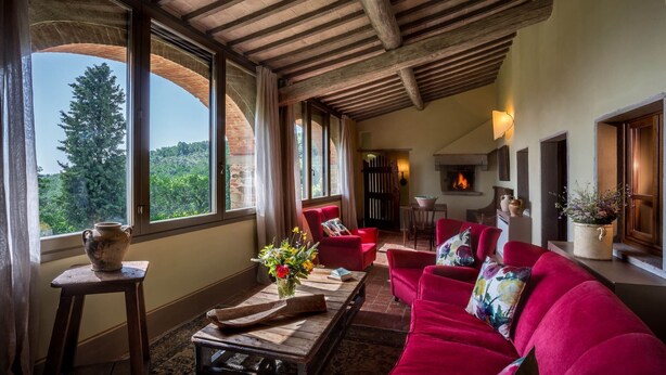 A cozy living room with a red sofa, a wooden coffee table, and a fireplace. Large windows offer a view of the outdoors. - Podere della Rosa: elegance and comfort in the Tuscan hills (Arezzo)