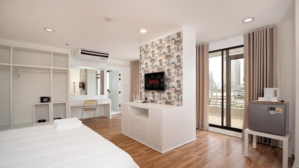 A modern hotel room with a large bed, a desk with a computer, a TV mounted on the wall, and a balcony with a view of the city. - Bed to Bliss (Bangkok)