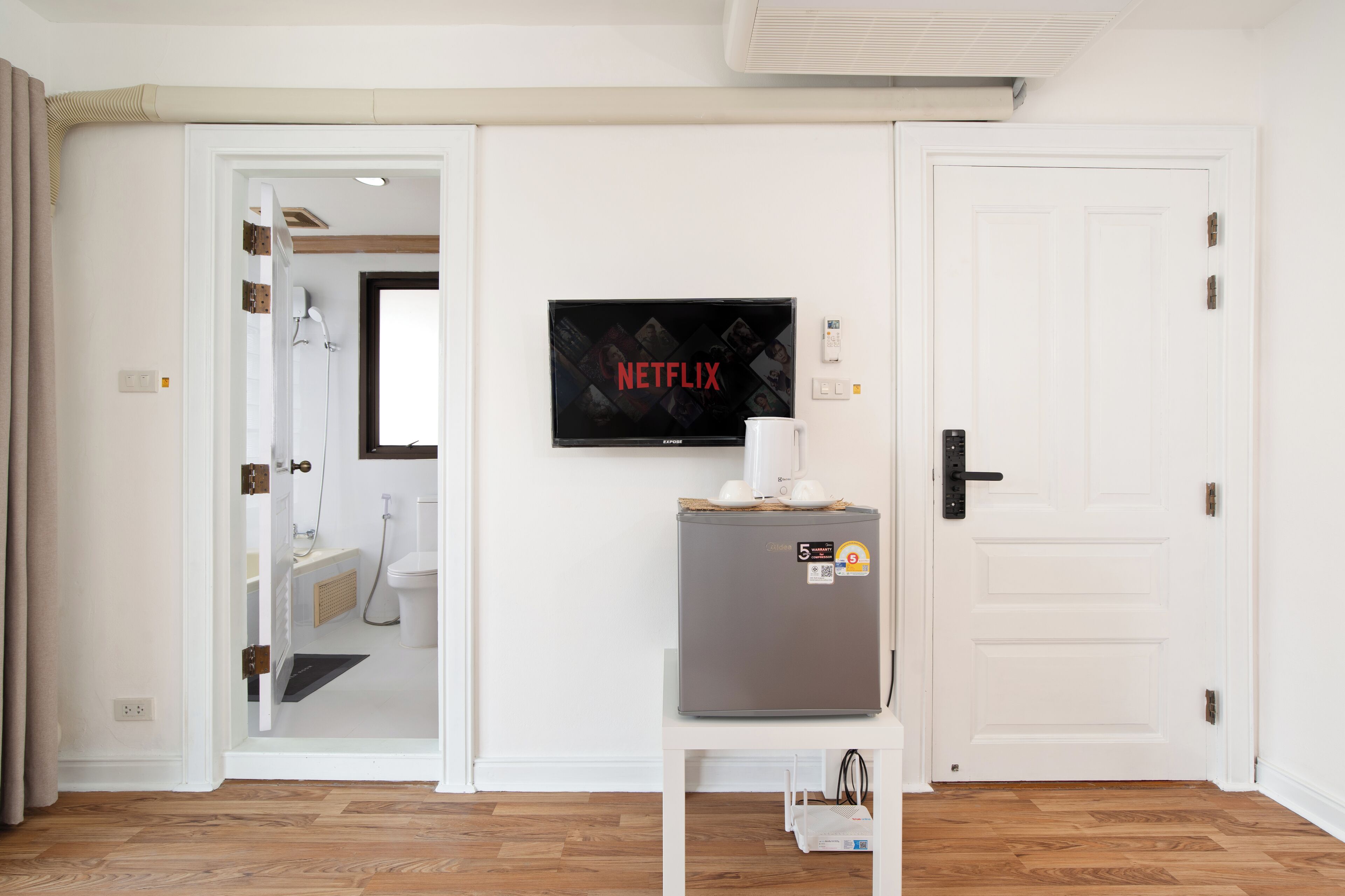 Ein Zimmer mit einer weißen Tür, einem an der Wand befestigten Fernseher, der 'NETFLIX' zeigt, einem kleinen Tisch mit einer weißen Tasse und einem grauen Kühlschrank.