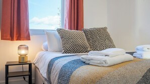 Una cama bien hecha con almohadas estampadas, una lámpara de noche y toallas dobladas en una mesita de noche junto a una ventana con cortinas.
