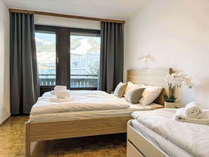 Ein Hotelzimmer mit einem großen Bett, einem kleineren Bett, einem Nachttisch, einer Wandlampe und einem Fenster mit Vorhängen.