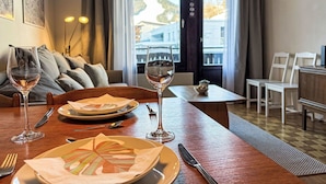 Et hotelværelse med et spisebord dækket til to personer, to vinglas, en tallerken med serviet og en lille skål på bordet. Der er en sofa med puder og et vindue med gardiner.