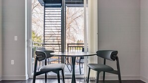 Habitació moderna amb taula rodona i dues cadires, porta corredissa de vidre que dona a un balcó, i vista a arbres i un edifici a l'exterior.