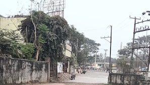 A wide, empty road with a concrete surface and patches of dirt. There are trees and a building with a sign on the left. - Aryan (Bhubaneshwar)