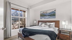 Un dormitorio con una cama grande, una ventana con cortinas, un cuadro de montañas y una mesita de noche con una lámpara.