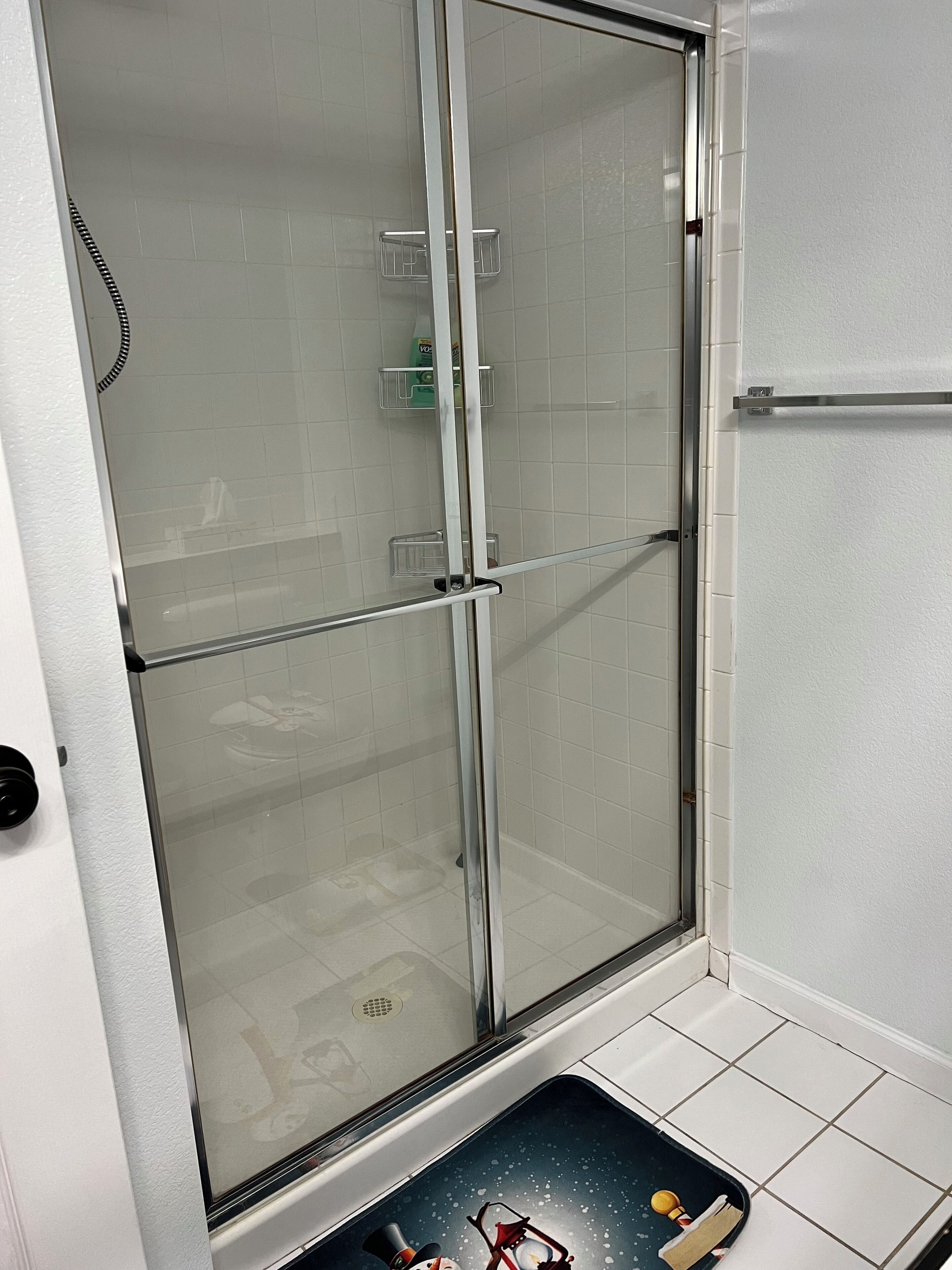 A glass-enclosed shower stall with a white tiled wall, a metal towel rack, and a small shelf with toiletries.