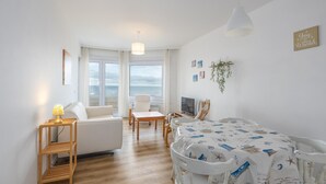 Una habitación luminosa y espaciosa con un sofá blanco, una mesa de comedor con mantel de temática náutica y vistas al mar a través de grandes ventanales.