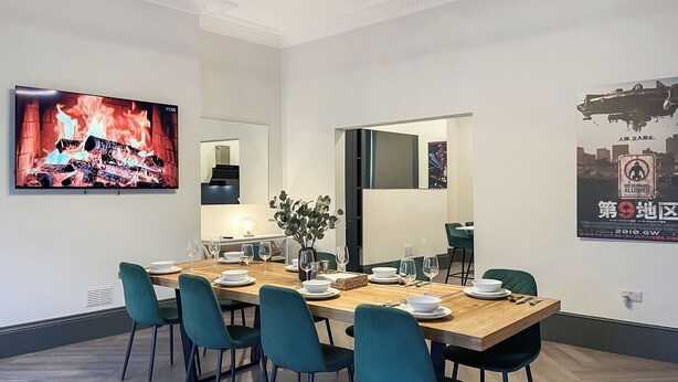 A modern dining area with a wooden table, teal chairs, and a flat-screen TV displaying a fire scene.