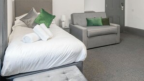 A hotel room with a bed, a grey sofa, a small table with a lamp, and a door.