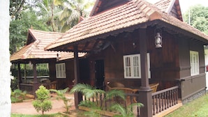 A traditional wooden house with a tiled roof, a porch with a railing, and a small garden with palm trees.