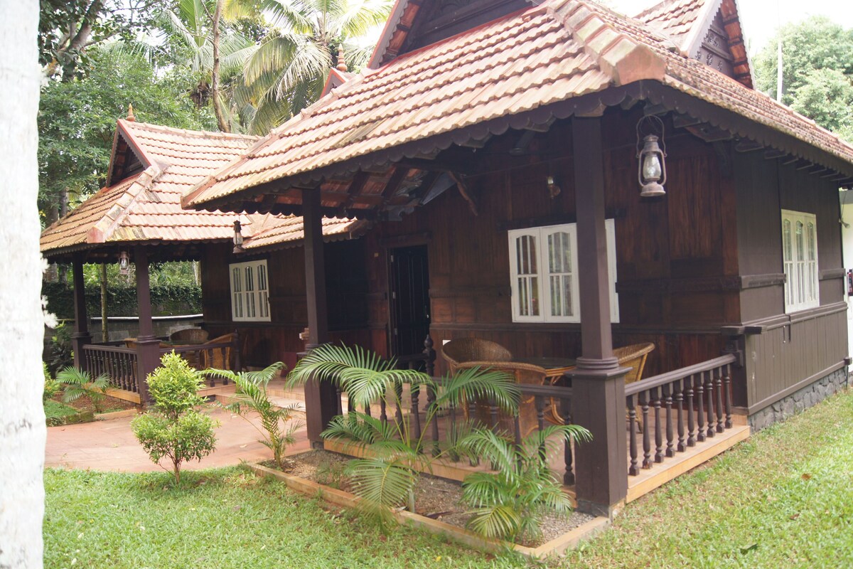 A traditional wooden house with a tiled roof, a porch with a railing, and a small garden with palm trees.