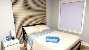 Un lit bien fait, avec une serviette bleue, une table de chevet sur laquelle se trouvent des bouteilles d’eau, et un téléviseur fixé au mur.