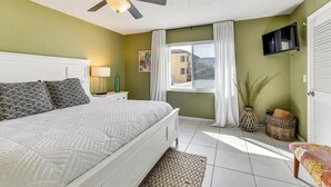 A bedroom with a bed, a ceiling fan, a TV mounted on the wall, a window with curtains, and a view of a building outside.