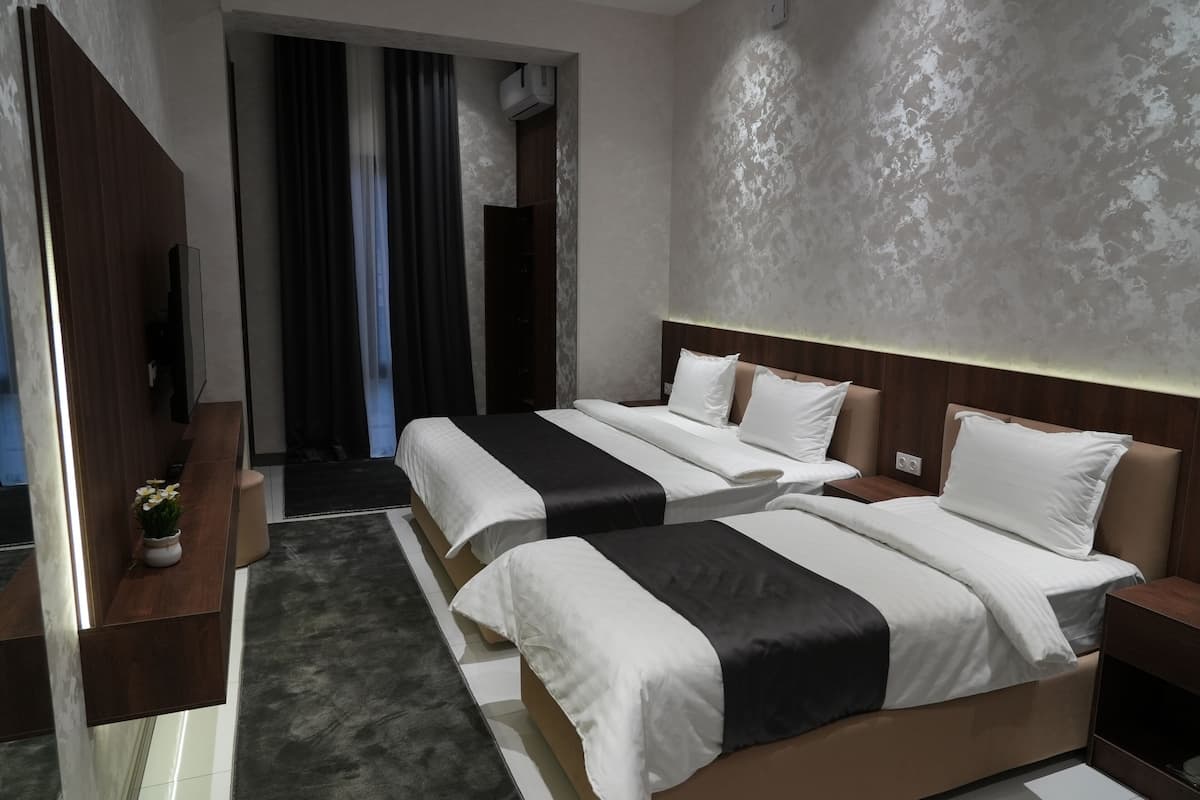 Ein Hotelzimmer mit zwei Betten, einem Schreibtisch und einem Fernseher. Ein Fenster mit Vorhängen und eine Tür zu einem Flur sind ebenfalls vorhanden.