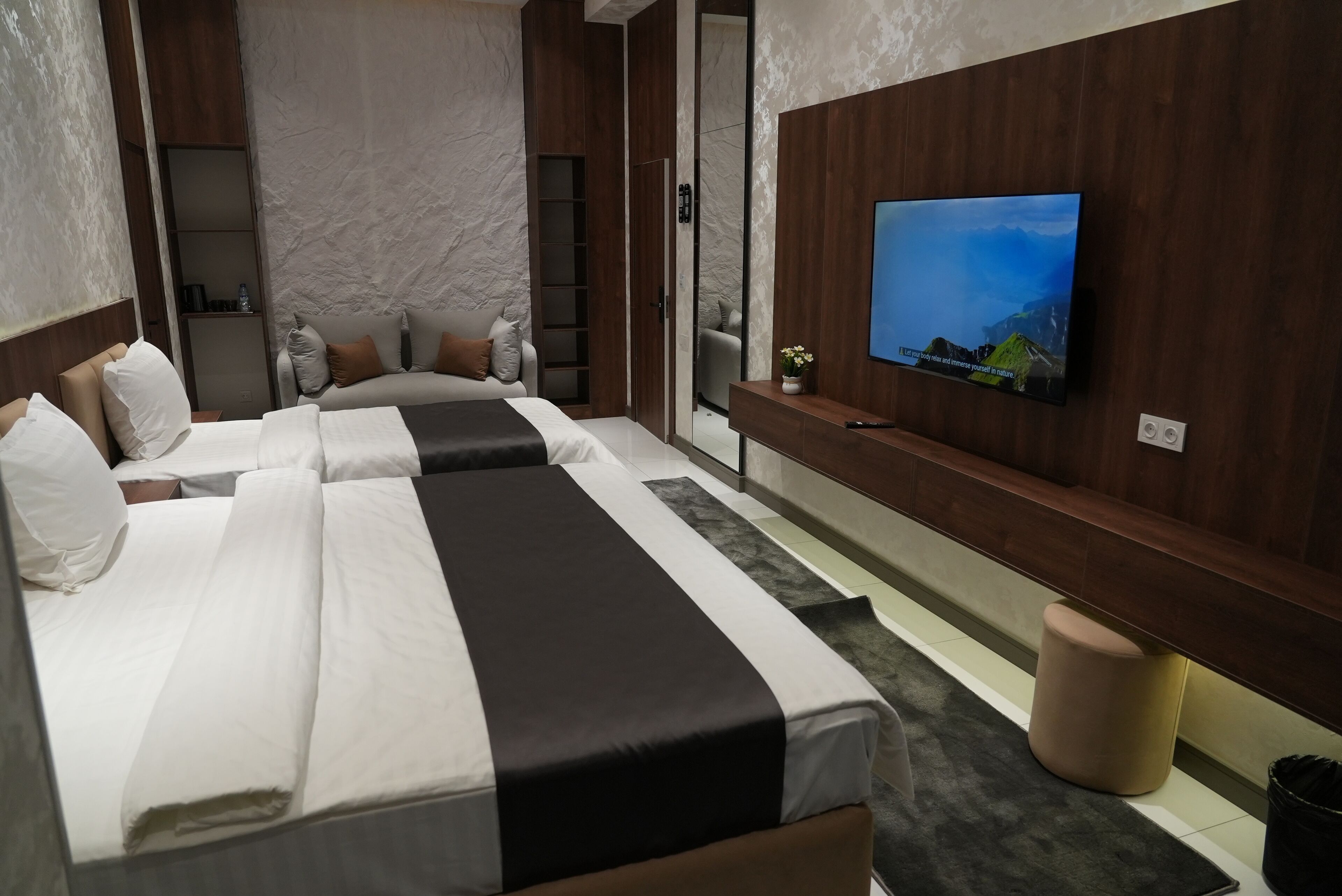 A modern hotel room with two beds, a TV on the wall, and a small seating area.