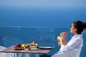 A person seated at a table with a variety of food items and a drink, overlooking a body of water. - Lilium Hotel Santorini (Santorini)