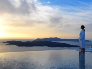 A person in a white robe standing on a balcony overlooking a body of water with distant landforms under a partly cloudy sky. - Lilium Hotel Santorini (Santorini)