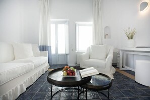 Un salon moderne avec un canapé blanc, un fauteuil blanc et deux tables rondes, dont l’une est ornée d’un panier de fruits et d’un livre.