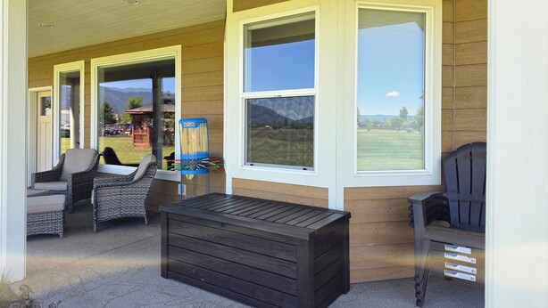 A covered porch with wicker furniture, a dark wooden bench, and large windows offering a view of a green landscape. - Wheelchair Accessible - Sleeps 10 - Mountain Views - Near Glacier Nat'l Park  (Kalispell)
