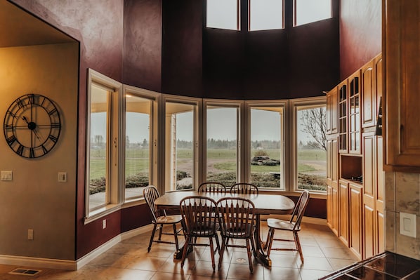 A dining area with a large table and chairs, large windows with a view of the outdoors, and wooden cabinetry.