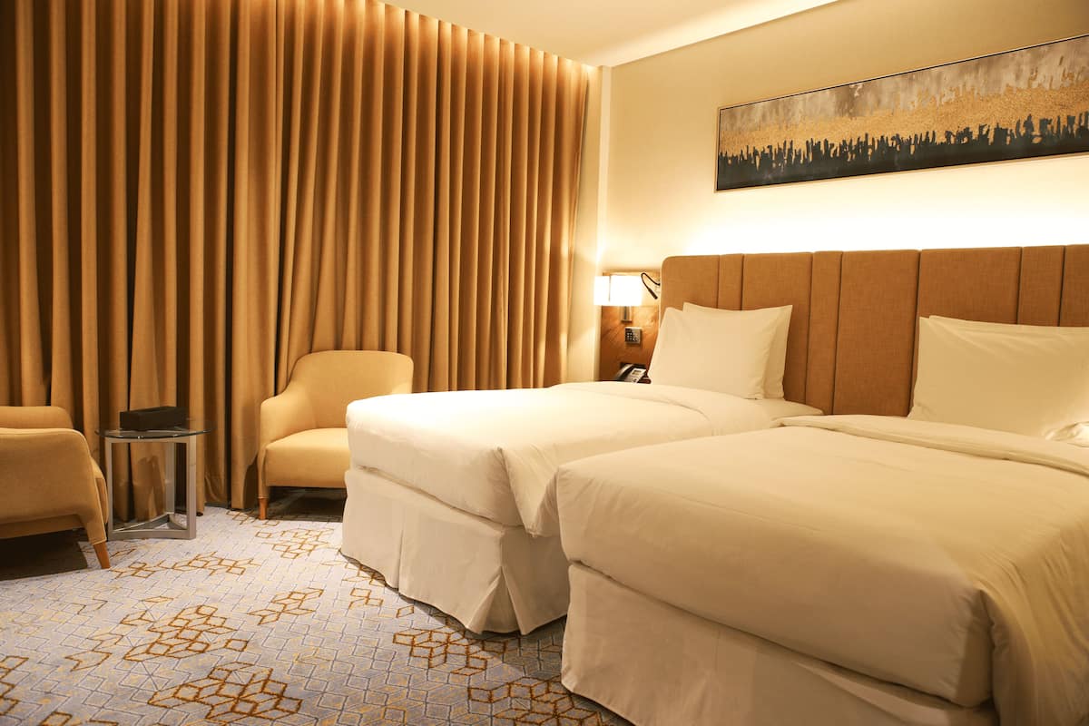 Habitación de hotel con dos camas, un área de descanso con una silla, una mesita y un cuadro en la pared.