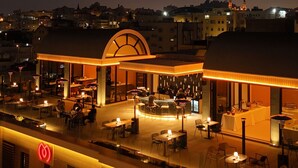 A hotel with a prominent arch entrance, outdoor seating area, and a visible sign with Arabic script.