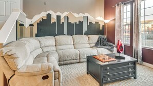 Ein Wohnzimmer mit einem großen, L-förmigen Ecksofa, einem schwarzen Couchtisch mit Schachbrettmuster und einer Wanddekoration mit Stadtansicht.