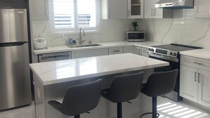 A modern kitchen with white cabinets, a stainless steel refrigerator, a built-in oven, and a central island with bar stools. - Brand-New Modern 2BR /2BA Private Home – Near Little Saigon, K-Town & Disneyland (Santa Ana)
