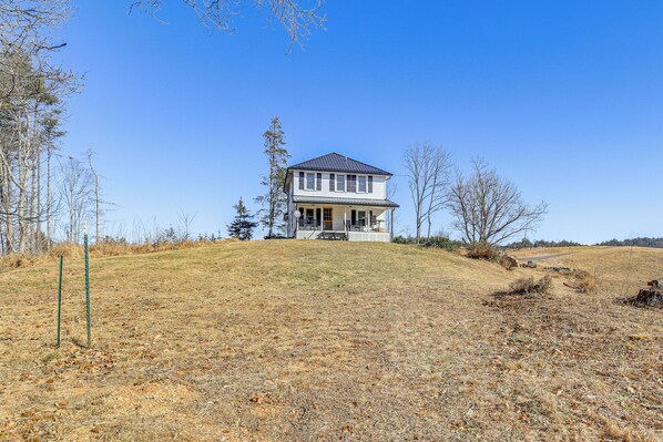 A two-story house with a front porch, situated on a grassy hill with trees around. - Mtn Views, New River Access & 98 Acres: Galax Gem (Galax)