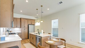 A modern kitchen with wooden cabinets, a stainless steel refrigerator, and a dining area with a round table and chairs. - Near NRG Stadium & Med Center | Group Retreat (Houston)