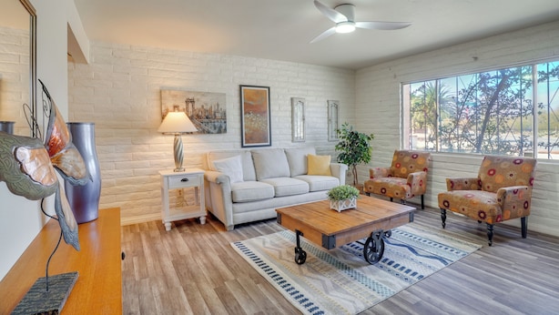 A spacious living room with a white brick wall, a ceiling fan, a large window with a view, a sofa, armchairs, a coffee table, and a side table with a lamp. - Hike, Golf & Dine: Tucson Retreat w/ Mtn Views! (Tucson)
